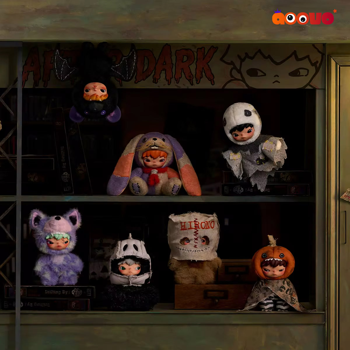 Six cute, stylized character plush toys dressed in spooky Halloween costumes are displayed on two shelves inside a dark, atmospheric shop window. The costumes include a black cat with orange eyes, a purple bunny with orange ears, a gray ghost, a purple cat with green eyes, a black skull-faced figure, a white bandaged head with red text, and a pumpkin-headed skeleton.