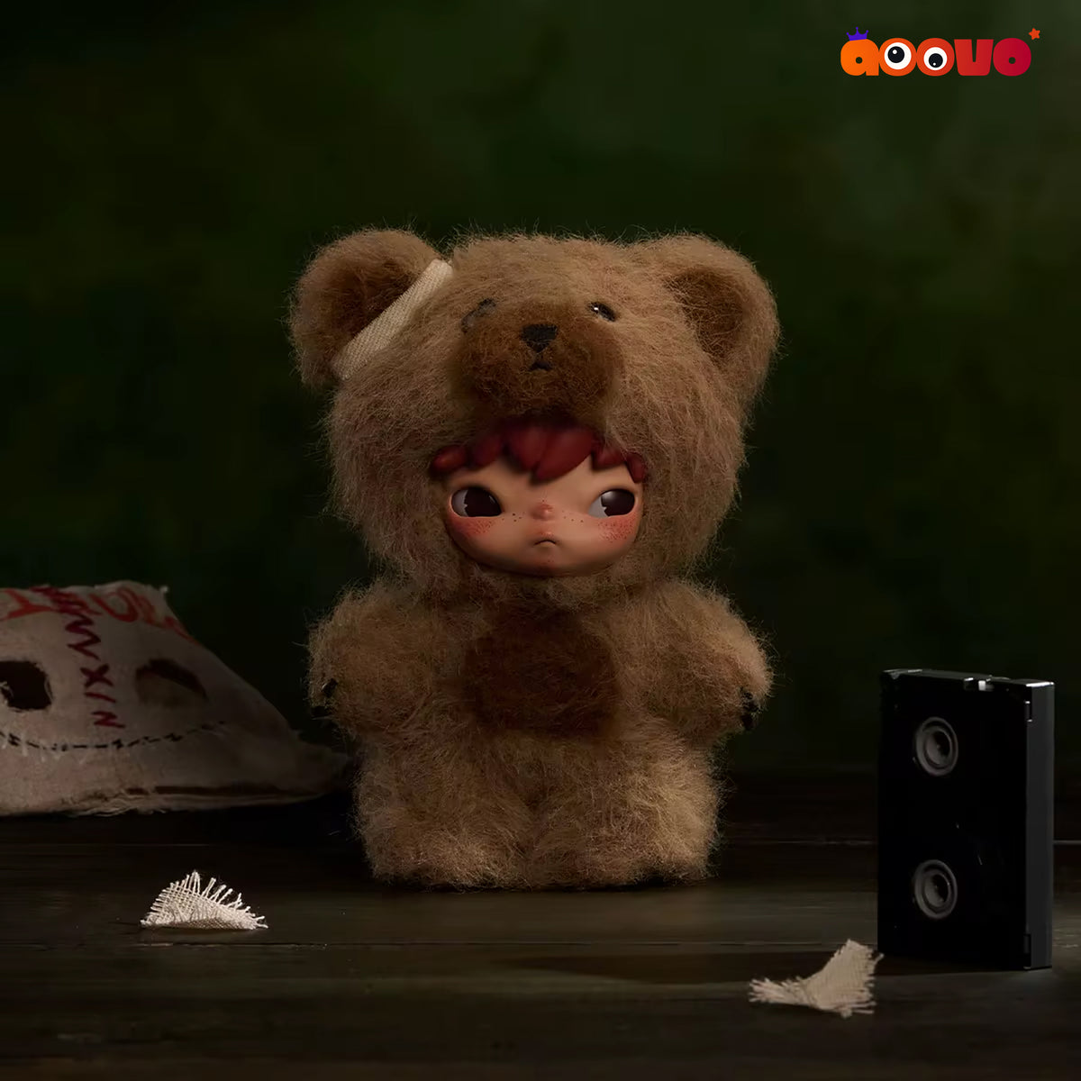 A small doll with large eyes and red hair is dressed in a fuzzy brown teddy bear costume, standing on a dark surface against a dark green background. A black speaker, a torn mask, and white shreds are placed around it.