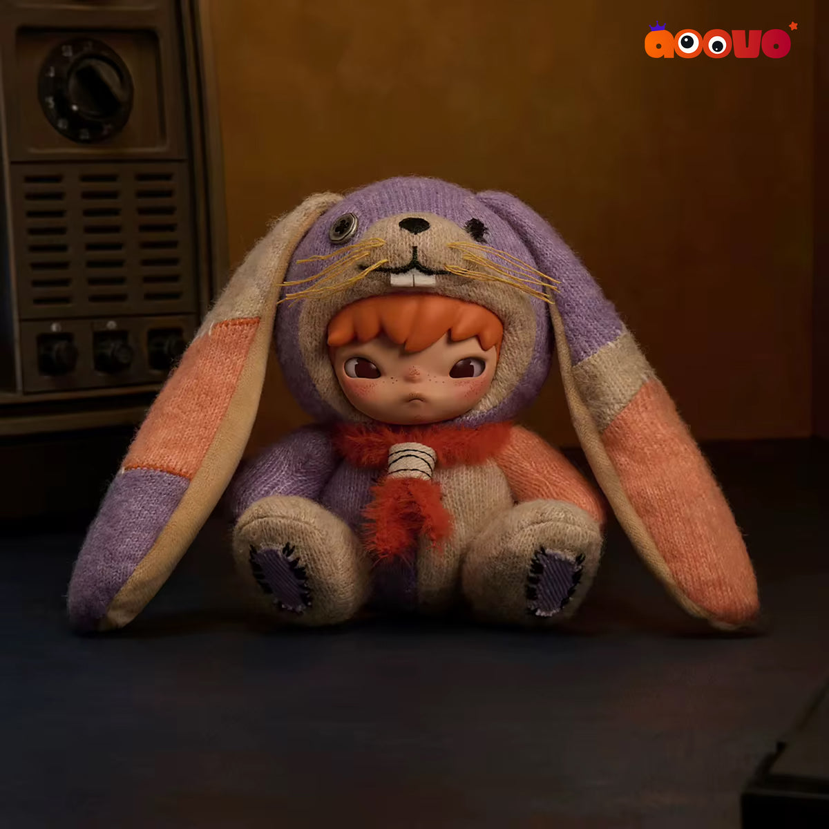 A plush doll dressed in a knitted bunny costume, sitting on a dark surface in a dimly lit room with a vintage radio in the background. The bunny costume is lavender purple with peach and beige patches, and the doll has orange hair and a red scarf.