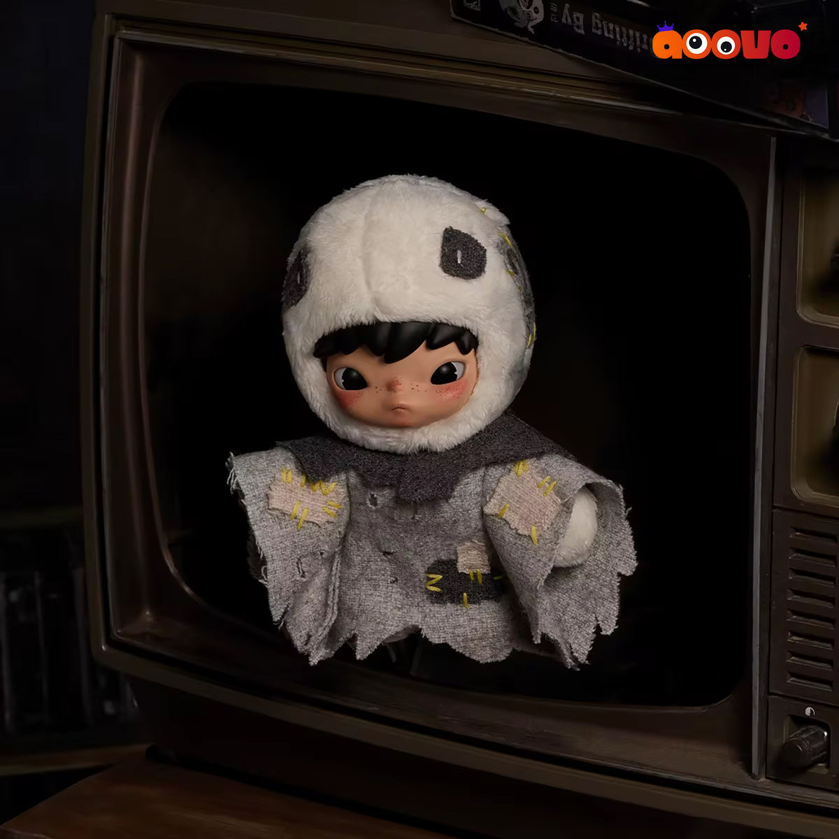 A cute doll with large eyes, black hair, and a fuzzy white hood sits inside the dark screen of a vintage brown television. The doll wears a gray tattered cape with yellow embroidered details.