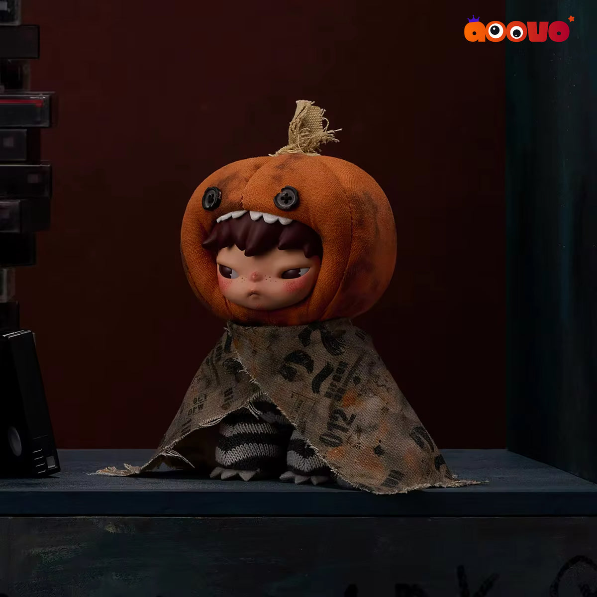 A stylized collectible doll or figurine wearing a carved orange pumpkin head with stitched facial features and a tattered brown-and-black patterned cloak, posed on a dark tabletop against a moody dark red and blue background.