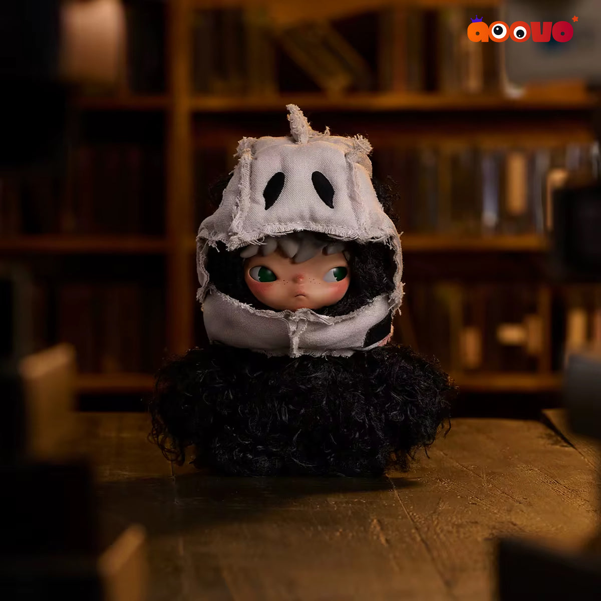 A doll or collectible figure with large green eyes and pale skin peeking out from a tattered white hood, wearing a fluffy black outfit, posed on a wooden table in a dimly lit library or study with bookshelves in the background.