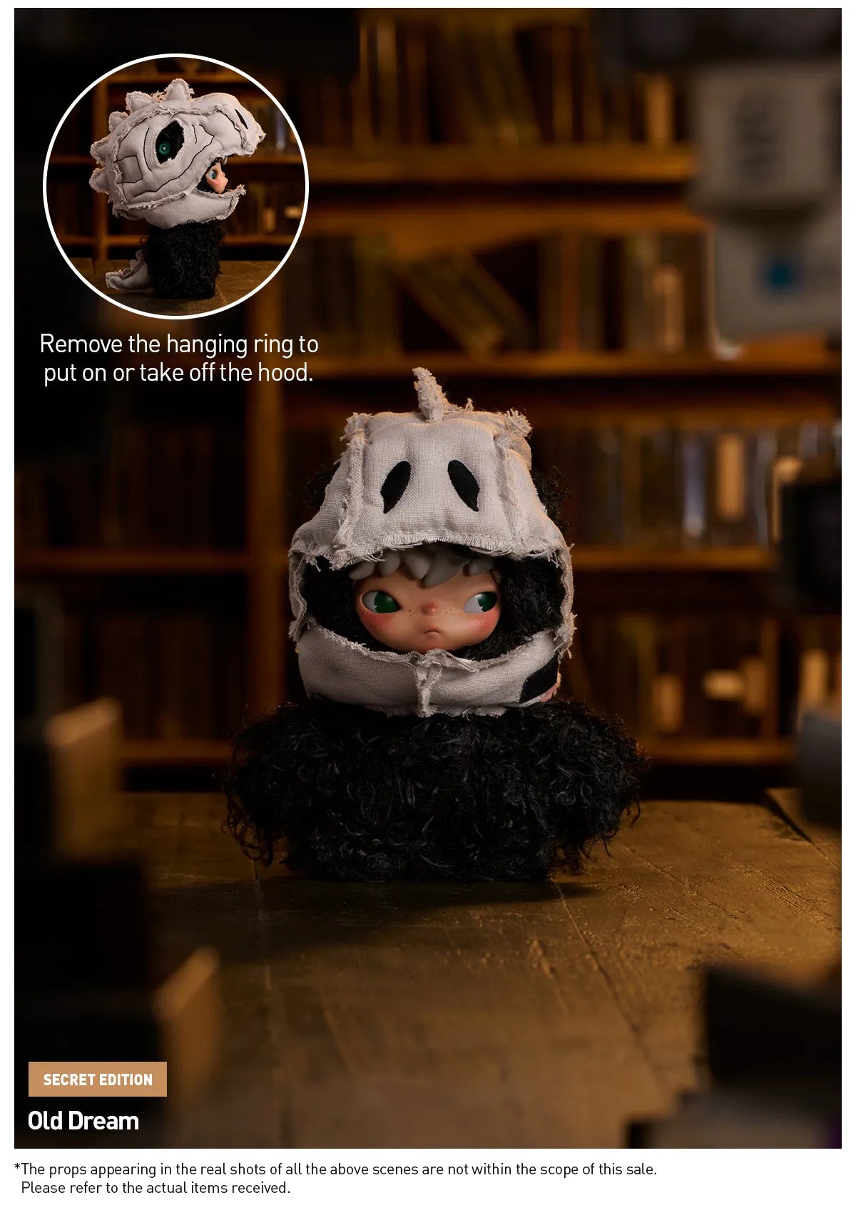 A doll or collectible figure wearing a black fluffy outfit and a white hood with black eye cutouts, standing on a wooden surface in a dimly lit room with bookshelves in the background. A circular inset at the top shows the hood being removed, with text explaining how to put on or take off the hood.